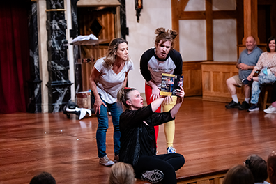 Jenny Bennett sitting on the front of the stage and wearing black shirt and pants holds up the play program  for Ginna Hoben in white t-shirt and geans, and Allie Babich in black and gray t-shirt ad red and yellow tights, both leaning over to read what Bennett is pointing at in the program